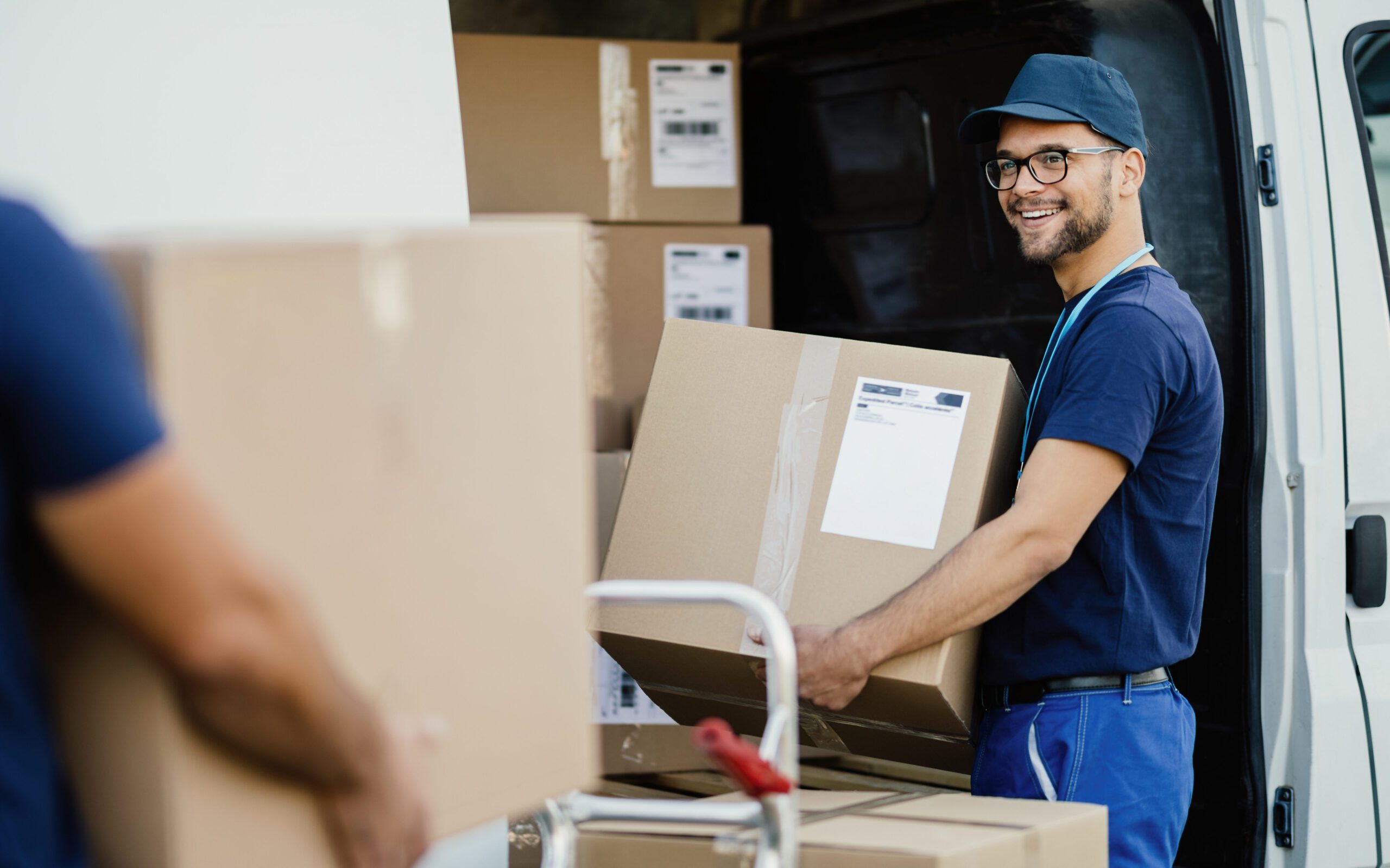 Young happy manual worker carrying cardboard boxes in the delivery van while communicating with his colleagues.