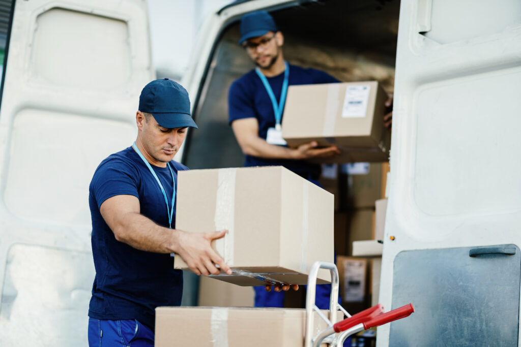 Young courier and his colleague unloading cardboard boxes from the delivery van.